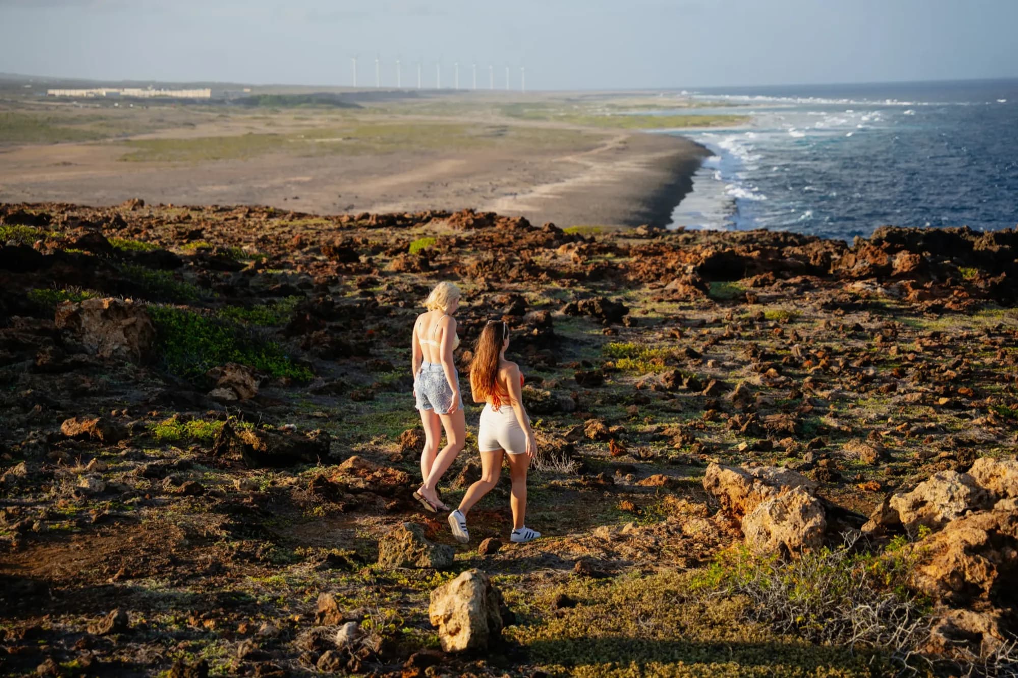 Two women walking along a rocky black-rock coastline at golden hour in Aruba