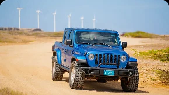 Front three-quarter view of a blue Jeep Gladiator pickup on a dirt road with wind turbines behind it in Aruba