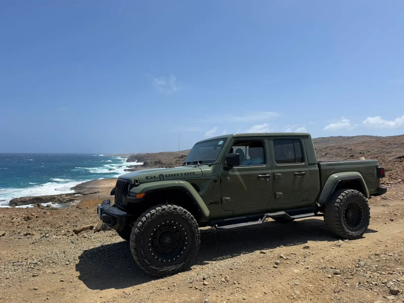 An olive green Jeep Gladiator parked on a rocky cliff edge overlooking the ocean in Aruba