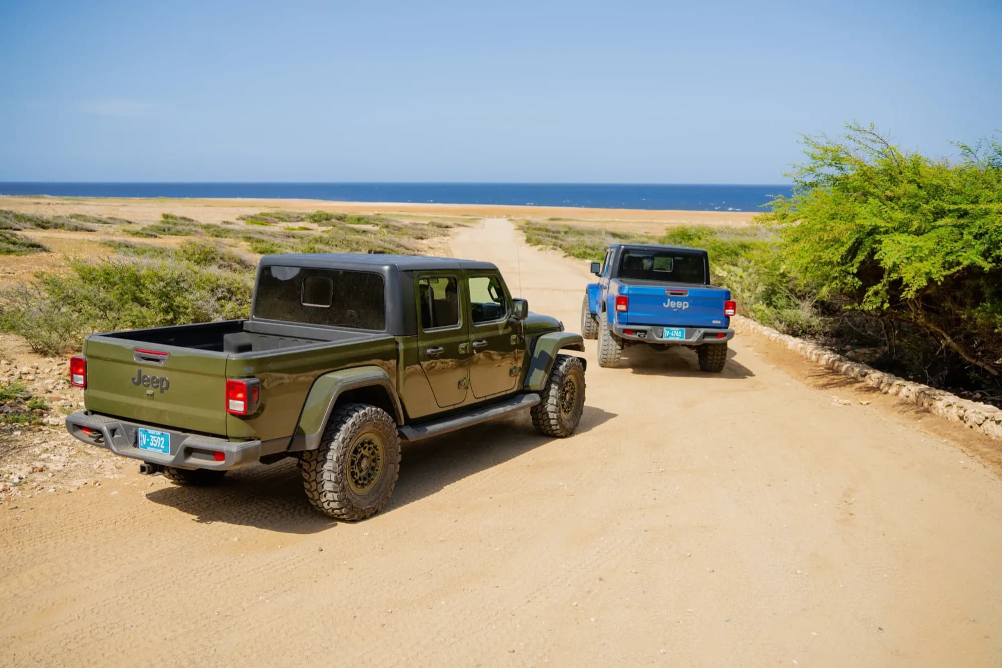 Two Jeep pickups seen from behind on a dirt path lined with a low stone wall along the Aruba coast