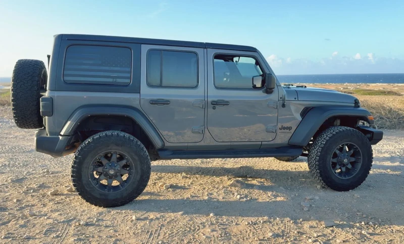 Side profile of a gray Jeep Wrangler parked on a dirt path with the ocean behind it in Aruba