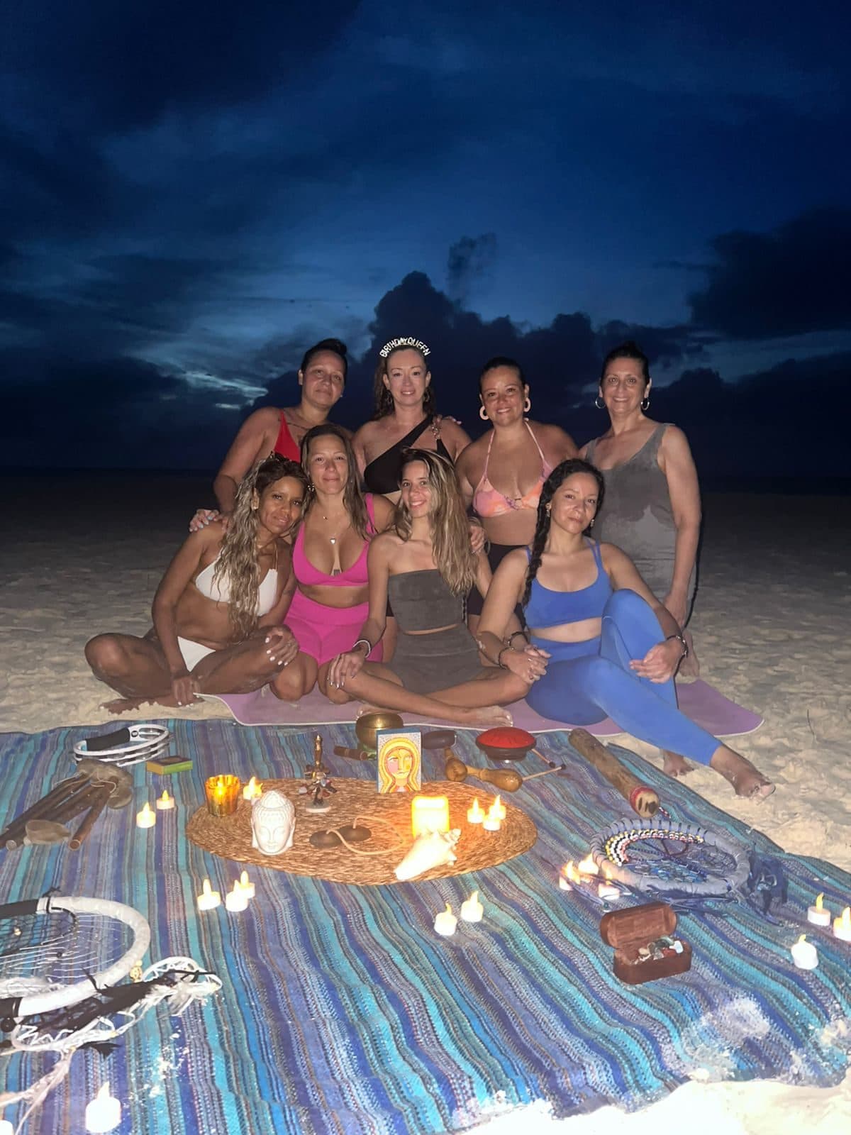 A smiling group gathered around candlelight on the beach after an evening sound healing session