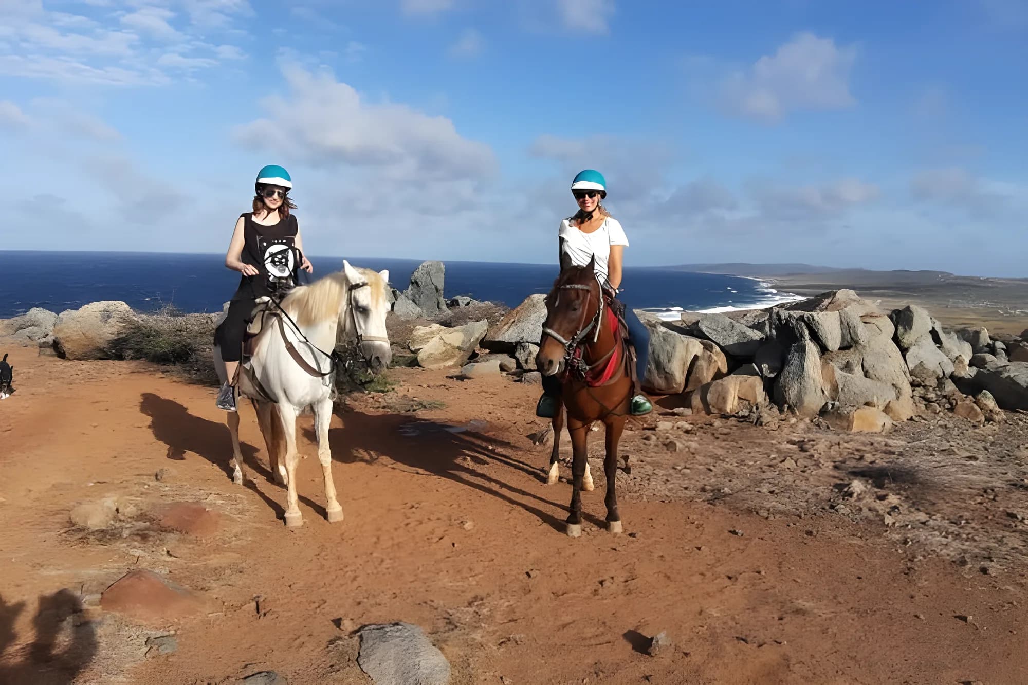 Two riders in helmets on horseback on the rocky coast of Aruba
