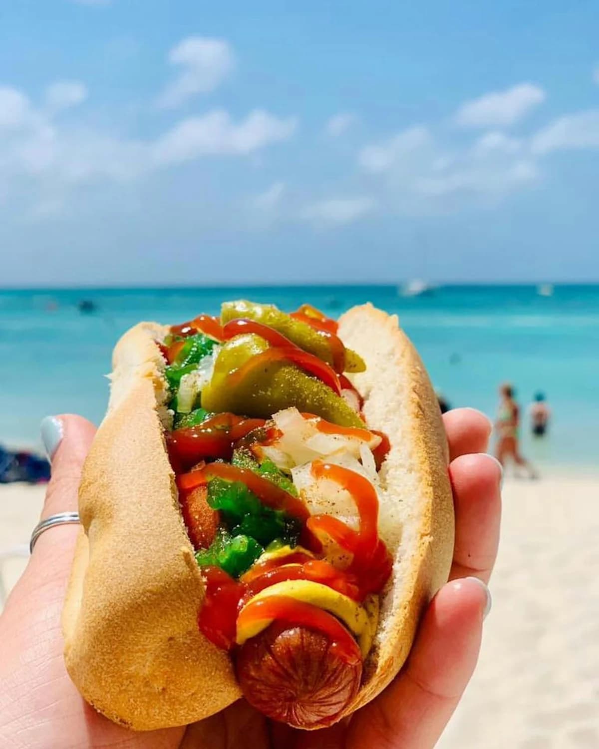 A topped bratwurst from Scott’s Brats held beside the turquoise water of Palm Beach, Aruba